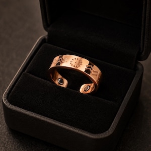 Könnte beinhalten: Ein kupferfarbener Metallring mit offenem Banddesign, verziert mit eingravierten Zickzacklinien und einem zentralen sonnenartigen Motiv. Die Innenseite des Rings zeigt zwei kleine, dunkle, kreisförmige Magneteinsätze in einer schwarzen Box.