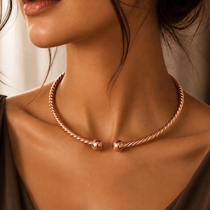 Könnte beinhalten: Ein roségoldfarbenes Choker-Halsband mit einem gedrehten, seilartigen Metallband. Das Design ist vorne offen und endet in zwei glatten, polierten kugelförmigen Endstücken, die sich in der Mitte des Halses gegenüberstehen.