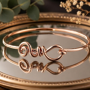 Puede incluir: Un brazalete de metal en tono oro rosa con un elemento decorativo central compuesto por un alambre continuo formado en una serie de bucles y espirales interconectados, creando un diseño fluido y abstracto sobre una banda circular fina y pulida.