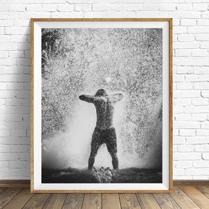 May include: A black and white photograph of a person standing in a shower of water droplets. The person is facing away from the camera with their arms raised above their head. The water droplets are creating a blurry effect around the person.