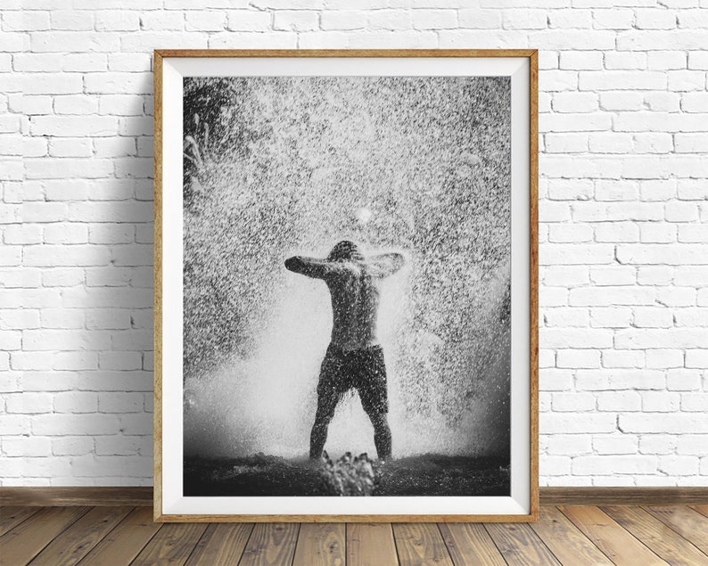May include: A black and white photograph of a person standing in a shower of water droplets. The person is facing away from the camera with their arms raised above their head. The water droplets are creating a blurry effect around the person.