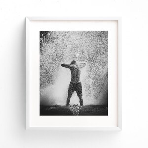 May include: A black and white photograph of a person standing in a fountain with water splashing around them.