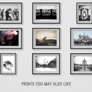May include: A wall gallery of nine black and white framed photographs. The photos include a group of people dancing, a subway car interior, a brick archway, a person holding a red flare, a surfer on a wave, a view of Notre Dame Cathedral in Paris, a tree with a cloudy background, a snowy landscape with a church, and a view of a building with a dome.