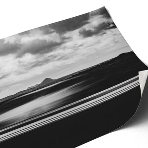 Photographie en noir et blanc capturant un paysage serein. L’avant-plan présente une voiture blanche en filé de vitesse. Au milieu, on aperçoit des silhouettes de montagnes sous un vaste ciel strié de nuages.