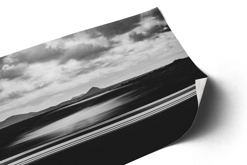 Photographie en noir et blanc capturant un paysage serein. L’avant-plan présente une voiture blanche en filé de vitesse. Au milieu, on aperçoit des silhouettes de montagnes sous un vaste ciel strié de nuages.