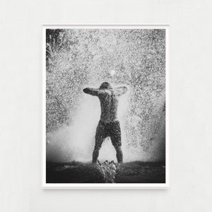 May include: A person stands with arms raised, facing away from the camera, as water splashes around them. The image is in black and white.