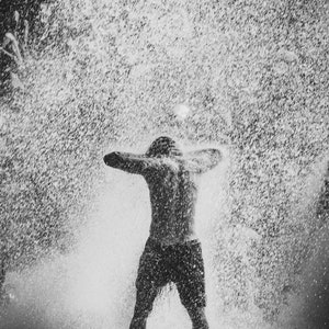 May include: A person standing in a shower of water with their arms raised above their head. The water is spraying from a sprinkler or other source, creating a misty effect.