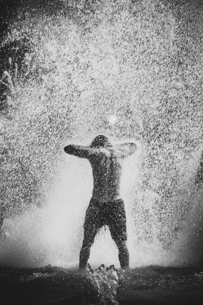 May include: A person standing in a shower of water with their arms raised above their head. The water is spraying from a sprinkler or other source, creating a misty effect.