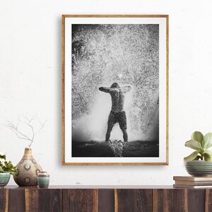 May include: Black and white photograph of a person standing in a shower of water droplets. The person is facing away from the camera with their arms raised above their head.