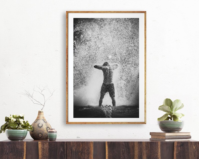 May include: Black and white photograph of a person standing in a shower of water droplets. The person is facing away from the camera with their arms raised above their head.