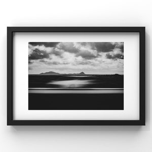 Photographie en noir et blanc capturant un paysage serein. L’avant-plan présente une voiture blanche en filé de vitesse. Au milieu, on aperçoit des silhouettes de montagnes sous un vaste ciel strié de nuages.