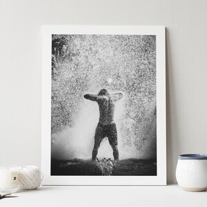 May include: A black and white photograph of a person standing in a fountain with water spraying up around them.