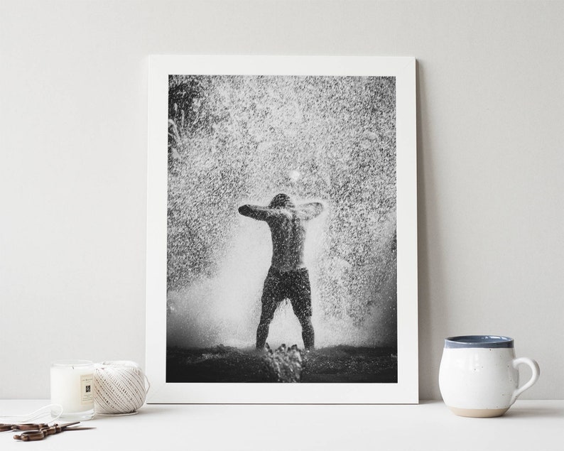 May include: A black and white photograph of a person standing in a fountain with water spraying up around them.