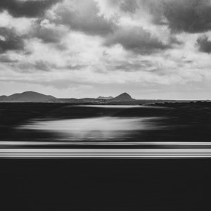 Photographie en noir et blanc capturant un paysage serein. L’avant-plan présente une voiture blanche en filé de vitesse. Au milieu, on aperçoit des silhouettes de montagnes sous un vaste ciel strié de nuages.