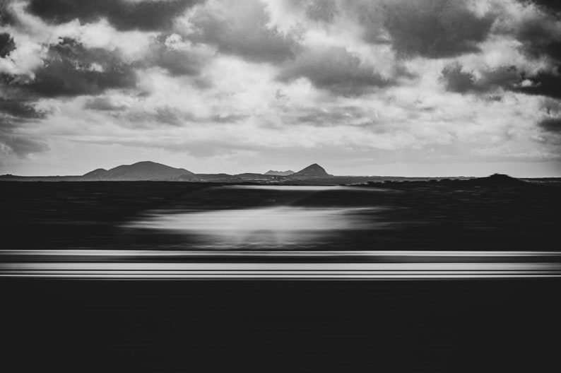 Photographie en noir et blanc capturant un paysage serein. L’avant-plan présente une voiture blanche en filé de vitesse. Au milieu, on aperçoit des silhouettes de montagnes sous un vaste ciel strié de nuages.