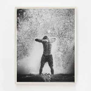May include: A black and white photograph of a person standing in a shower of water droplets. The person is facing away from the camera and has their arms raised above their head. The water droplets are falling all around them.
