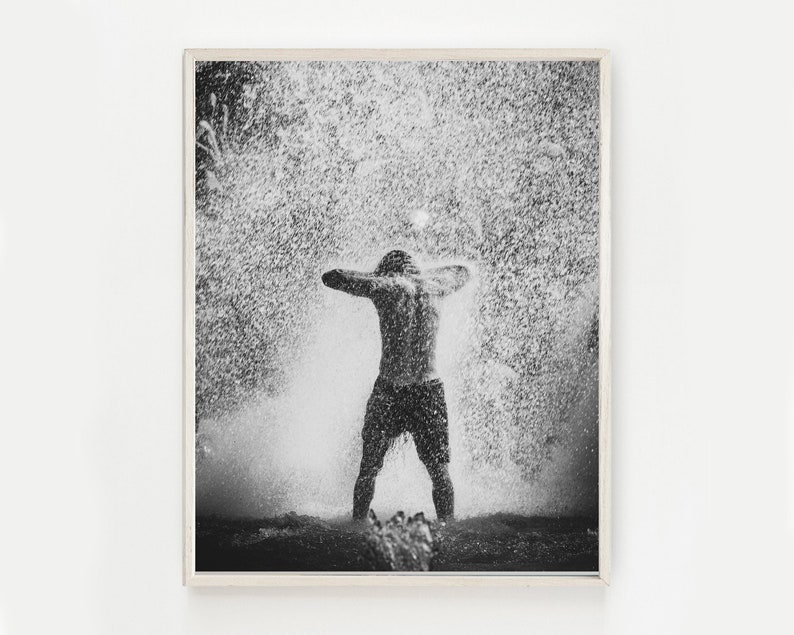 May include: A black and white photograph of a person standing in a shower of water droplets. The person is facing away from the camera and has their arms raised above their head. The water droplets are falling all around them.