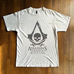 Pode incluir: Uma t-shirt branca de manga curta com um grande gráfico cinzento no peito. O design apresenta o logótipo de Assassin's Creed com um crânio e espadas cruzadas, acompanhado pelo texto "ASSASSIN'S CREED IV BLACK FLAG" impresso abaixo.