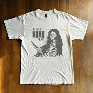 Può includere: Una t-shirt bianca a maniche corte con una stampa grafica grigia di uno scheletro che tiene in mano un coltello. Il testo riporta BLACK FLAG e LIVE AT THE ON BROADWAY 23 JULY 1982. Il design include quattro barre verticali.