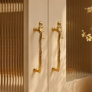 May include: Two gold-coloured metal cabinet handles mounted on off-white cabinet doors with vertical reeded glass panels. Each handle features a textured branch-like stem topped with a small bird figure, providing a decorative grip for the doors.