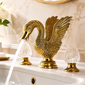 May include: A gold-coloured swan-shaped bathroom tap with detailed feather textures and wings extended upwards. The tap features two clear, textured crystal-like handles on gold bases, with water flowing from the swan's beak into a white sink.