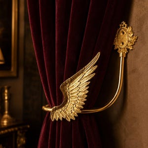 May include: A gold-colored metal curtain tieback featuring a detailed wing design. The curved arm extends from a decorative circular wall-mounted plate, holding a deep burgundy velvet curtain in place with the wing element facing forward.