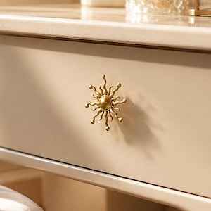 May include: A cream-colored vanity table featuring a single drawer with a gold sun-shaped knob. The tabletop holds a ribbed white vase with pink flowers, a clear textured glass candle holder, and a gold-framed jewelry tray with pearl beads.