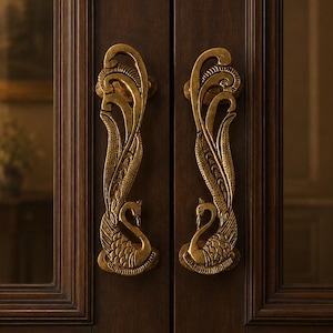 May include: A pair of vertical brass door handles mounted on dark brown wooden double doors. Each handle features an intricate, embossed design of a swan with long, curved, feathered wings extending upward along the length of the metal hardware.