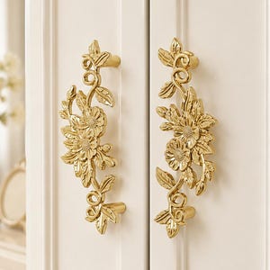 May include: Two gold-coloured metal cabinet handles mounted on a white panelled door. Each handle features a detailed relief design of flowers, leaves, and winding vines, with two attachment points securing the hardware to the door surface.