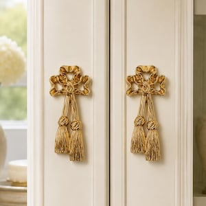 May include: Two gold-toned metal cabinet pulls shaped like decorative bows with hanging tassels. The pulls are mounted on a pair of off-white cabinet doors with vertical paneling, featuring a polished finish that reflects light on the curved surfaces.