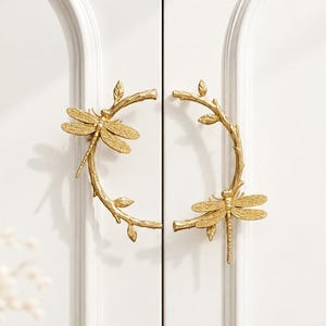 May include: A pair of gold-colored metal cabinet handles, each shaped like a curved branch with small leaves and a detailed dragonfly perched on the stem. The handles are mounted on white double doors with arched paneling and a smooth finish.