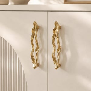 May include: Two vertical cabinet handles in a polished gold finish, featuring an irregular, organic, and wavy silhouette. These metal pulls are mounted onto a smooth, matte off-white cabinet door surface, creating a distinct contrast in texture and colour.