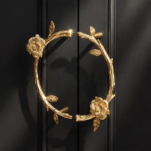 May include: A pair of gold-colored metal cabinet handles shaped like curved branches with textured bark, small leaves, and a prominent blooming flower on each handle. The handles are mounted vertically on a matte black double-door cabinet surface.