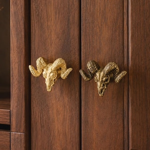 Könnte beinhalten: Zwei Schrankknöpfe aus Metall in Form von Widderschädeln, die an einer Schranktür aus dunklem Holz befestigt sind. Der linke Knopf hat ein glänzendes Gold-Finish, während der rechte Knopf ein dunkleres, antikes Messing-Finish aufweist.