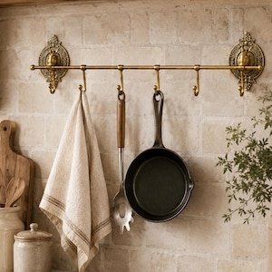 Puede incluir: Un riel de cocina de latón con soportes de pared ornamentados y cinco ganchos. Colgados de los ganchos hay un paño de cocina de lino beige, una cuchara de servir de acero inoxidable con mango de madera y una sartén de hierro fundido negra.