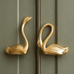 May include: Two gold-coloured metal cabinet pulls shaped like swans. One swan is depicted with its neck extended upwards, while the other swan is shown with its neck curved downwards. Both pieces feature detailed, textured wing patterns.