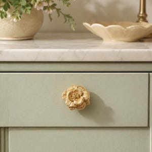 May include: A gold-colored metal cabinet knob shaped like a blooming flower with layered, textured petals. The knob is mounted on a flat, light sage green wooden drawer front, featuring a detailed, realistic floral design with a metallic finish.