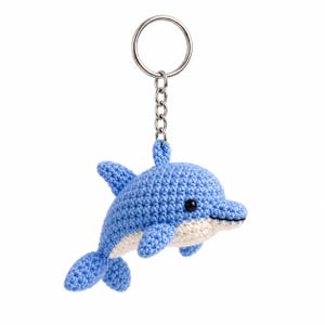 Può includere: Un portachiavi a forma di delfino all'uncinetto con il corpo superiore azzurro e il ventre color crema. Il delfino ha un piccolo occhio a perla nera ed è fissato a un anello metallico argentato tramite una catenella, creando un accessorio compatto.