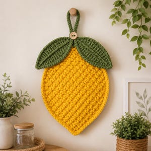 Puede incluir: Un agarrador de cocina de ganchillo con forma de limón, con un cuerpo texturizado de color amarillo brillante y dos hojas de color verde oscuro en la parte superior. Un pequeño botón de madera se sitúa donde se unen las hojas, con un lazo.