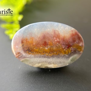 Puede incluir: Primer plano de un cabujón de ágata colorido con bandas rojas, naranjas, amarillas y blancas. La ágata tiene forma ovalada y una superficie lisa y pulida.