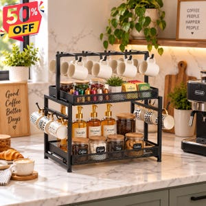 May include: A two-tier black metal coffee station organizer featuring a mesh tray, hooks for hanging mugs, and glass jars with labels for coffee, sugar, tea, and cookies. The unit holds syrup bottles and various mugs on a marble countertop.