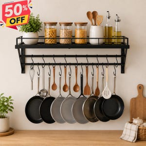 May include: A black metal wall-mounted kitchen storage rack with a top shelf holding three glass jars with wooden lids, a white ceramic utensil holder, and a green plant. S-hooks hang from the rack, supporting various metal pans and wooden spoons.