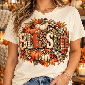 May include: A cream-colored short-sleeve t-shirt featuring the word BLESSED in large, patterned letters. The design is surrounded by orange and white pumpkins, autumn leaves, red berries, wheat stalks, and a green and white checkered ribbon bow.
