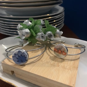 Peut inclure: Deux bracelets manchettes en métal argenté avec des inserts en céramique circulaires. Un insert présente un motif floral bleu, tandis que l'autre arbore un motif botanique orange rougeâtre. Les bracelets ont une structure à double fil.