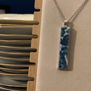 Può includere: Una collana con pendente rettangolare caratterizzata da un motivo floreale blu e bianco su una superficie simile alla ceramica, incastonata in una cornice di metallo argentato e sospesa a una sottile catenina argentata con motivi di foglie e petali.