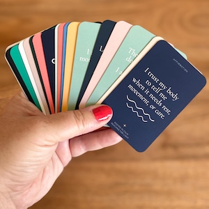 May include: A deck of rectangular affirmation cards fanned out, featuring a variety of solid colors including navy blue, mint green, soft pink, yellow, and coral. The top navy card displays the text: I trust my body to tell me when it needs rest, movement, or care.