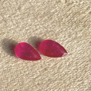 以下が含まれることがあります： 鮮やかで半透明なマゼンタピンクの色合いをした、ペアシェイプのルースストーン2個。それぞれの石は光を捉えるファセット加工された表面を持ち、尖った端と丸みを帯びたベースを持つ、滑らかで曲線的なティアドロップ型のシルエットをしています。