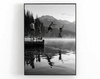 May include: A black and white framed wall art print depicting four individuals on a wooden dock at a lake. Two individuals stand on the dock while two others are captured mid-air jumping into the water. A forest and mountains are in the background.