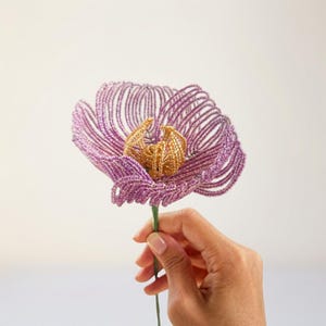 May include: A handcrafted flower sculpture constructed from small purple beads forming the petals and metallic gold beads forming the central stamen. The flower is attached to a thin green stem, creating a delicate and intricate botanical design.