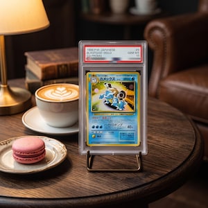 May include: A graded 1999 Japanese Blastoise Holo CD Promo trading card in a clear plastic slab. The card features a blue turtle character with cannons, Japanese text, and a Gem Mint 10 rating label at the top. The slab stands on a metal display stand.